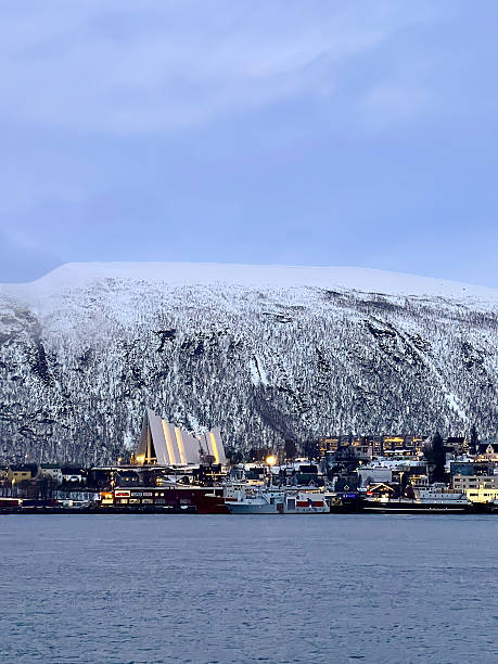 Tromso, Norway - December 22, 2025: Winter polar night. Daylight above the mainland mountain and its agglomeration with the cathedral on the left. Line of docked ships.