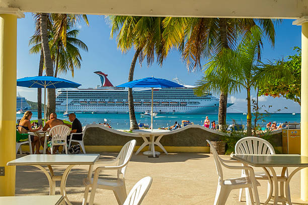 Ocho Rios, Jamaica - December 30, 2019: People enjoying lunch at an outdoor restaurant with a large cruise ship docked in the clear blue Caribbean sea, surrounded by palm trees and beachgoers
