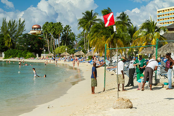 Ocho Rios, Jamaica - December 30, 2019: Tourists enjoying a sunny day at a tropical beach with palm trees, clear blue water, sunbathers on lounge chairs, and local vendors near a fence, showcasing Caribbean tourism
