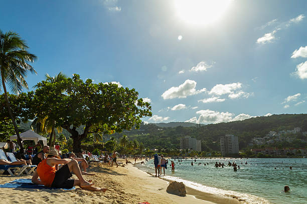 Ocho Rios, Jamaica - December 30, 2019: People are relaxing under palm trees and swimming in the clear blue sea on a sandy beach, enjoying a sunny day at a tropical tourist resort with lush green hills in the background