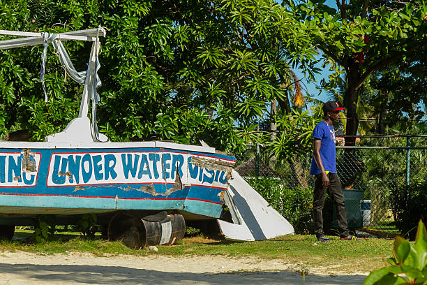 Ocho Rios, Jamaica - December 30, 2019: Young man walks past a weathered boat stenciled under water visual stranded on sandy shore beside green tropical trees under a bright blue Jamaican sky