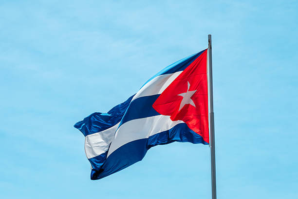 Cuban flag waving proudly on a tall flagpole against a bright blue sky. The bold red, white, and blue stripes create a dynamic, patriotic scene perfect for travel, culture, and national symbol themes. Taken in Santiago de Cuba.