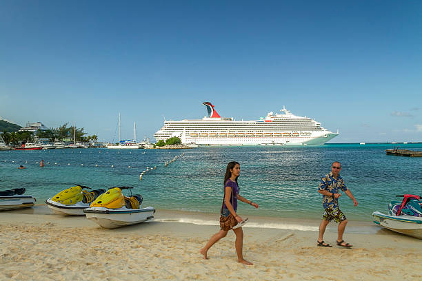Ocho Rios, Jamaica - December 30, 2019: Tourists stroll a sunny Ocho Rios beach beside turquoise Caribbean water with a large cruise ship docked in the bay, white sand, blue sky and tropical resort vibe