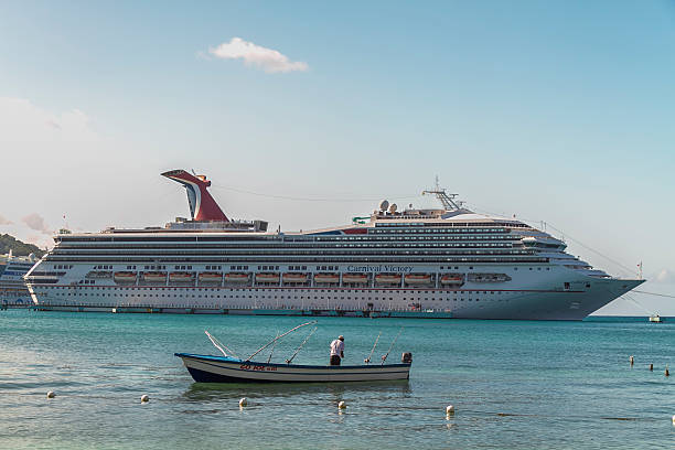 Ocho Rios, Jamaica - December 30, 2019: Fisherman standing on a small boat with fishing rods in the foreground, with the Carnival Victory cruise ship docked in the background and clear blue Caribbean waters