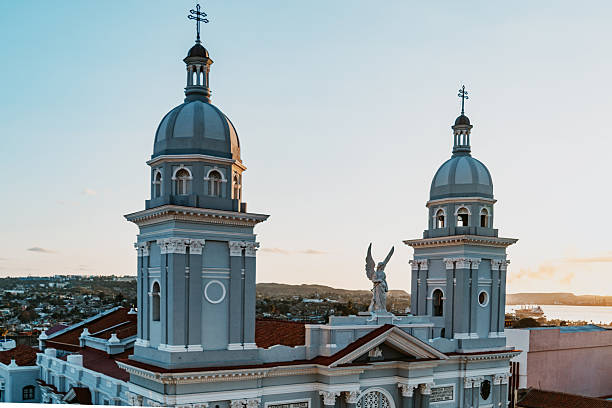 A grand church with twin domes rises above red-tiled roofs in Santiago de Cuba at golden hour. An angel statue guards the entrance, while pastel blue façades and a tranquil coastal backdrop evoke heritage.