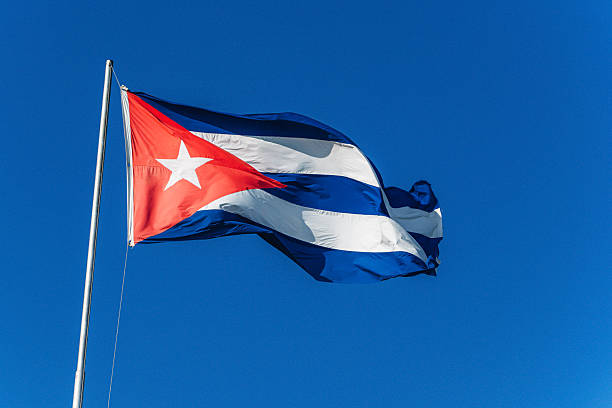 A vivid Cuban flag waves from a tall pole against a deep blue sky, symbolizing national pride, culture, and freedom. Captured outdoors in Santiago de Cuba, the image conveys energy and heritage.