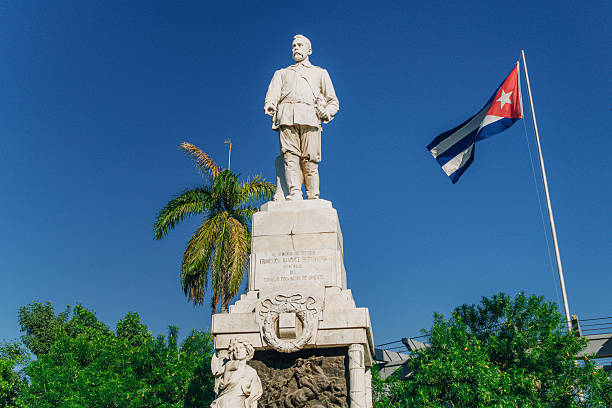 A tall stone statue stands atop a decorated pedestal beneath a clear blue sky, with a waving Cuban flag and lush tropical trees nearby, capturing national pride and historic monument vibes in Santiago de Cuba.