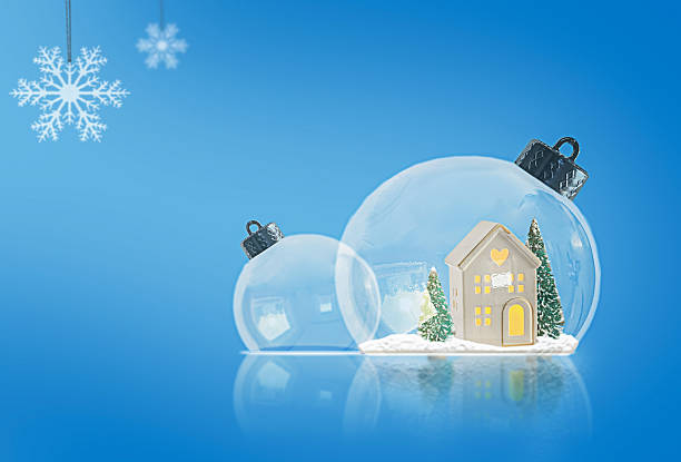 Two glass Christmas tree ornaments with miniature houses and trees inside. A smooth blue gradient background with snowflakes creates a festive atmosphere. High-quality photo.