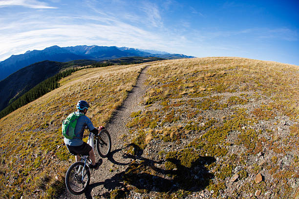 A man rides his modern, full-suspension, electric mountain bike down a singletrack trail in the Rocky Mountains of Alberta, Canada, on a sunny day. He wears cycling clothing, a backpack, and a bicycle helmet.