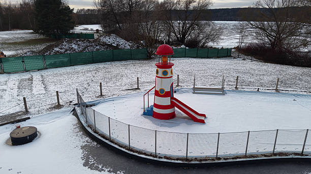 A red slide extends from the tower.   A fence surrounds the play area.   Trees and a river are visible in the background.   The scene appears wintry