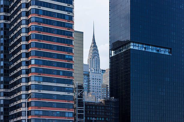 The famous Art Deco spire of the Chrysler Building is visible between two modern skyscrapers in Midtown Manhattan, New York City. On the right is a dark, glass-clad office building, and on the left is a residential tower with distinct horizontal bands of windows and panels. This close-up telephoto view highlights the architectural variety and density of the NYC skyline during a summer day.