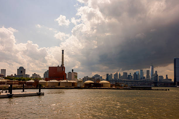 A view from a ferry on the East River in New York City on a summer afternoon. In the foreground, the industrial waterfront of Brooklyn features the East River Generating Station with its prominent smokestack and storage tanks. In the background, the iconic Lower Manhattan skyline, including One World Trade Center and the Manhattan Bridge, stands under a dramatic sky with dark, heavy storm clouds gathering on the right, contrasting with bright clouds on the left.