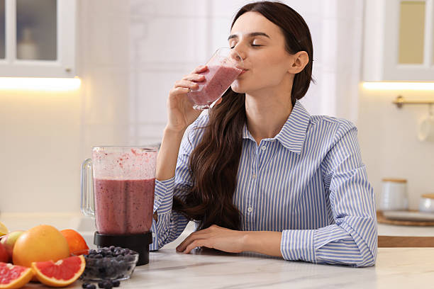 Woman drinking smoothie at white marble table with blender and fruits in kitchen