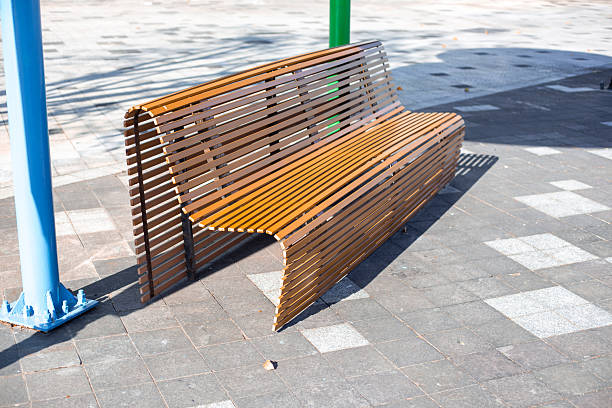A modern wooden bench with slats sits on a paved public square in bright sunlight next to colorful posts.
