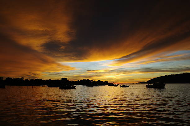 A stunning sunset over calm ocean water with dramatic golden and dark clouds stretching across the sky, while small boats appear as silhouettes on the horizon. The scene represents peace, adventure, travel, and natural beauty.