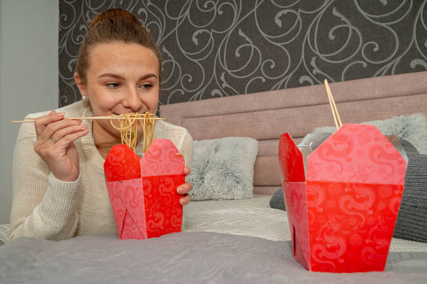 Two women enjoy eating takeaway on bed in bedroom. They are different ages. They eat food with chopsticks.