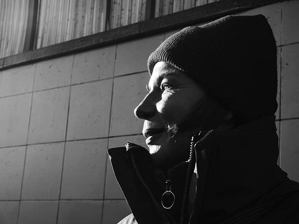 Person wearing a beanie and jacket smiling  in backlight , illuminated by dramatic side lighting against a tiled wall, creating a thoughtful and introspective mood