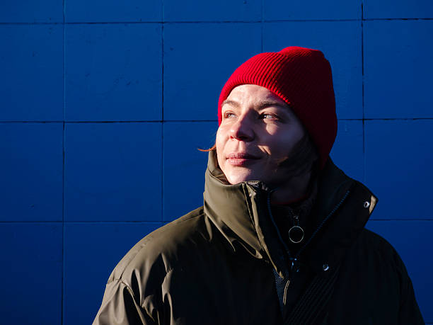 Woman standing outdoors, dressed in a warm coat and a red knit hat, looking confidently into the sun with natural skin against a vibrant blue wall background