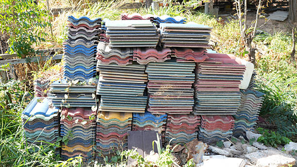Stacked roofing tiles in various colors create vibrant display in natural setting, surrounded by greenery and debris