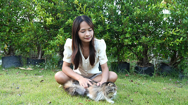 Woman sits on grass, gently petting fluffy cat in serene outdoor setting surrounded by greenery