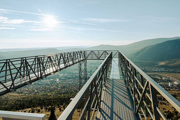 Glass skywalk walkway on mountain ridge with panoramic landscape and sunlight in background.
