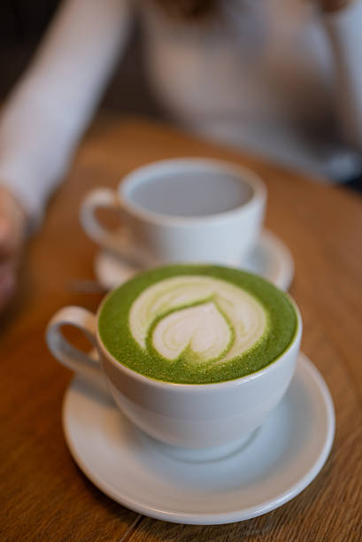 Creamy matcha latte forming heart-shaped art, nestled in white ceramic cup on rustic wooden surface, blurred coffee mug softening background ambiance