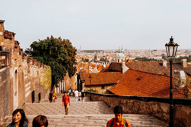 Prague, Czech Republic. August 21st, 2009: Tourists walking down a historic cobblestone path along ancient city walls with a panoramic view of Prague’s red rooftops and church domes in the background. Popular walking route combining historic architecture, tourism, and city life in the Czech capital. Editorial use only.