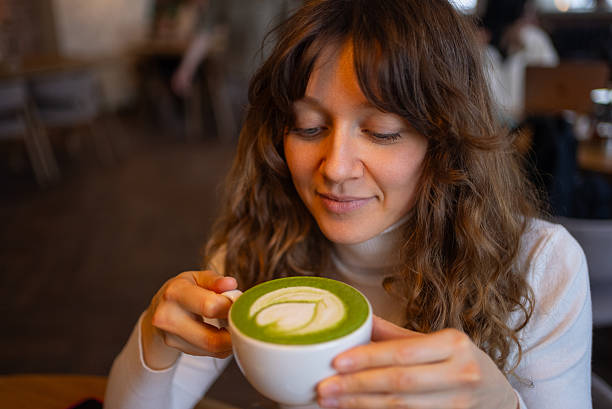 Young woman sipping matcha latte with delicate leaf art, relaxing in sunlit cafe corner, experiencing comforting green tea's smooth, earthy essence