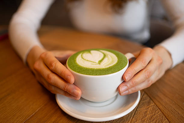 Feminine hands holding ceramic mug with matcha latte, delicate heart-shaped foam pattern visible, wooden surface background creating cozy coffee moment