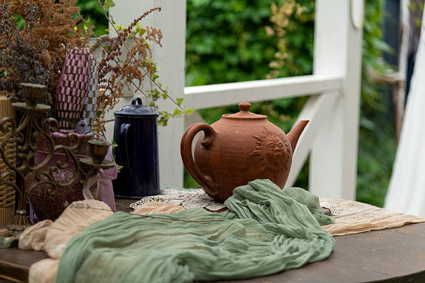 A teapot sits on a table next to a piece of cloth. There are dried plants and other items around it. The scene shows a green garden in the background with light filtering through.