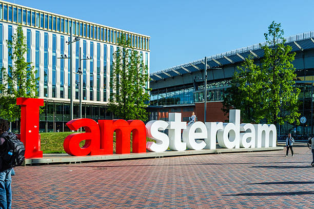 POV Point of view of latin backpacker tourist male holding a cardboard frame displaying the text I love Amsterdam, perfectly capturing a picturesque canal view adorned with iconic Dutch houses and boats under a cloudy sky
