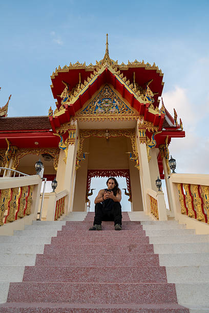 Man sitting on decorated temple stairs, looking at mobile phone, under a blue sky in thailand