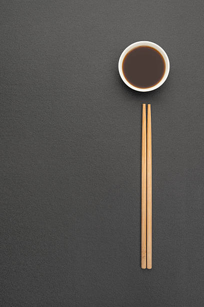 Black vertical textured background with white saucepan and soy sauce, with bamboo chopsticks