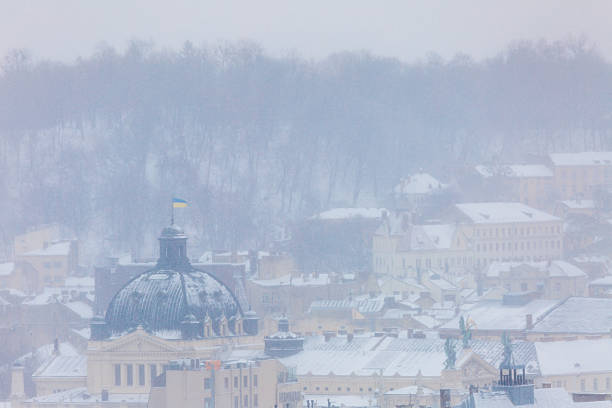Snow covered Lviv Opera House dome and Ivan Franko park during winter storm. Panoramic cityscape with historical landmarks and rooftops under heavy snowfall and blizzard on January day.
