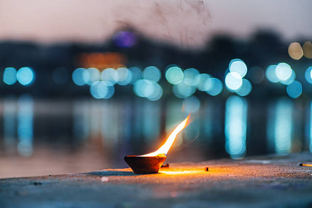 A single clay lamp burns on sandy ground beside a calm water body, with colorful city lights creating soft bokeh in the distance. Evokes warmth, spirituality, and evening serenity in Pushkar, India.