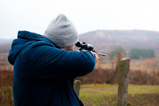 A skilled hunter stands in a vast open area, carefully aiming a rifle towards a far-off target. The scene captures a moment of focus amidst a cloudy sky.