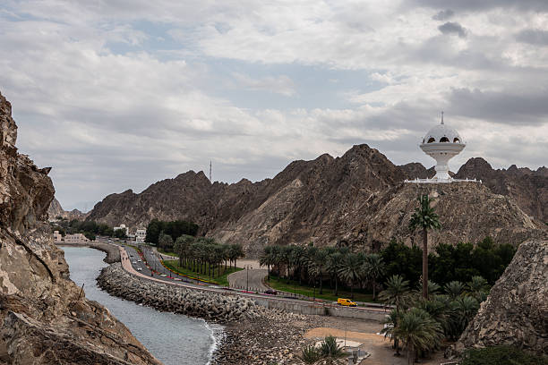 Sunset view of Muscat, featuring the tranquil waterfront, traditional architecture, and the historic Muttrah skyilne set against rugged mountains. A dhow peacefully floats in the Gulf of Oman, reflecting the city’s rich maritime heritage.