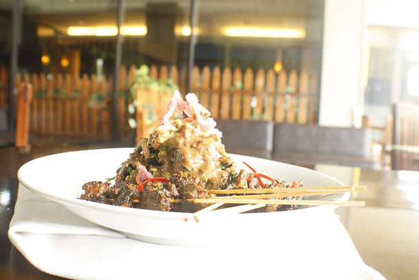 Close up of traditional Indonesian satay skewers topped with rich peanut sauce, bean sprouts, and red chili, served on a white plate in a restaurant