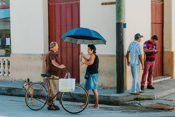 Gibara, Holguin Province, Cuba - February 13, 2017:  A lively Cuban street scene in Gibara shows an elderly man with a bicycle talking to a woman holding a blue umbrella while two men stand nearby. A dog accompanies the street life.