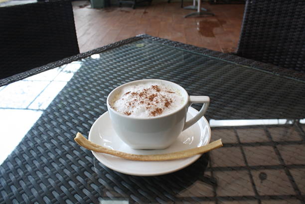 Warm cappuccino with foamed milk and a sprinkle of cinnamon, served with a delicate biscuit stick on a dark woven glass table in an outdoor cafe setting