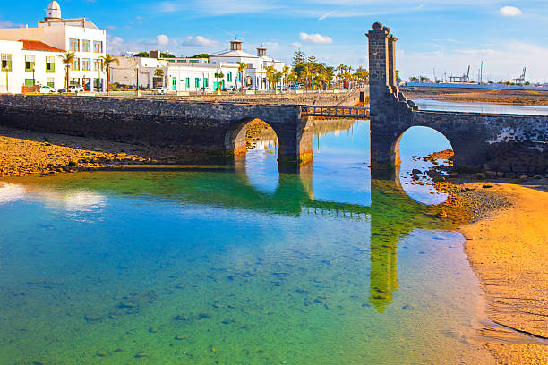 Puente de las Bolas in Arrecife, Lanzarote spans a calm lagoon with volcanic stone arches. Whitewashed buildings, palms, and blue skies reflect in clear water at low tide