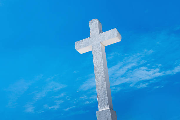 A tall white cross sculpture rises against a clear blue sky, symbolizing faith and spirituality. The textured surface catches bright sunlight, creating a striking contrast in Santiago de Cuba, Cuba.