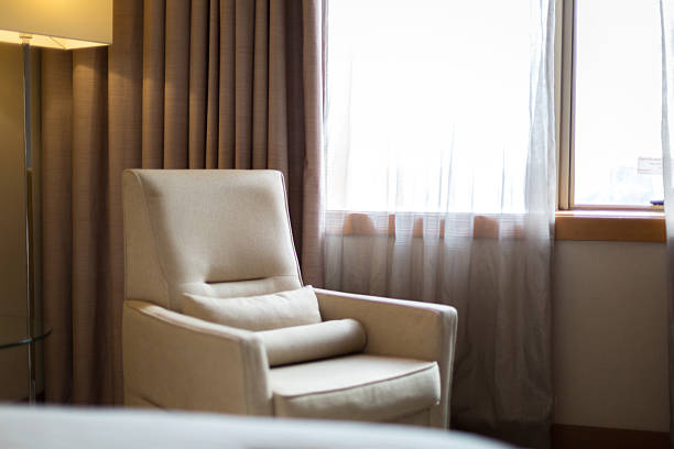 A calm and inviting hotel room interior featuring a comfortable armchair placed near a window with sheer curtains. Warm natural light fills the space, creating a cozy, home-like atmosphere ideal for relaxation, travel stays, or lifestyle concepts.
