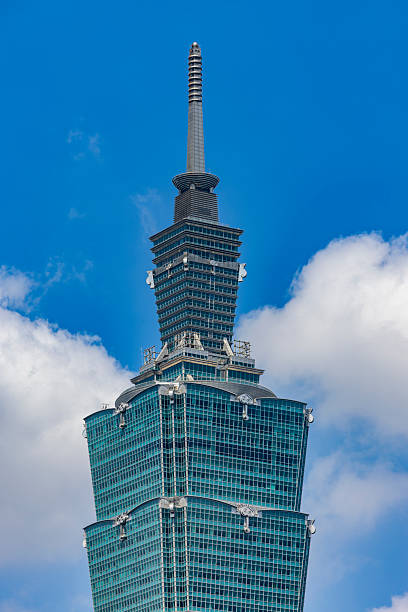 Close-up of the spire and upper tiers of Taipei 101 in Taipei, Taiwan. The iconic modern skyscraper is shown against a vivid blue sky with soft clouds, highlighting contemporary Asian architecture and a globally recognized urban landmark in daylight.