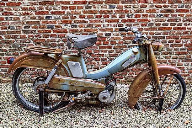 Classic motorcycle from 1960s, worn gold in color, street with small stones, brick wall in the background, old, broken, rusty and disused, old-fashioned display