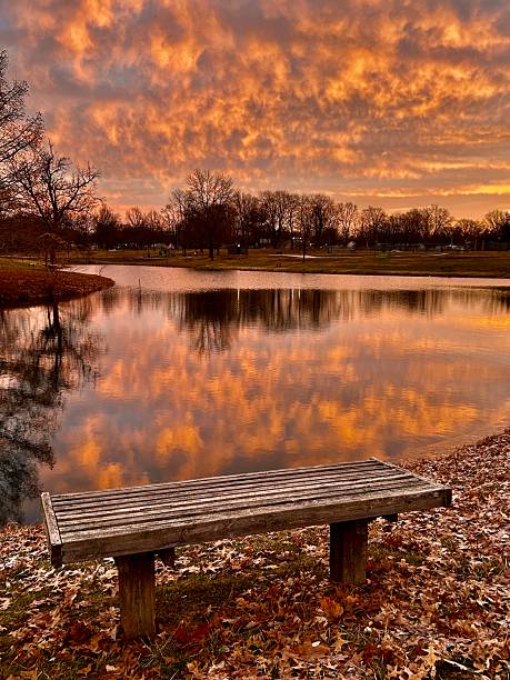 A cloudy sunrise in a park in the rural Midwest. A bench sits next to a lake as the sky turns pink and orange at dawn.