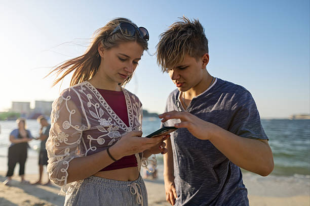 Two teens stand on a Dubai beach, heads tilted toward a phone as sunlight glints off sheer embroidery and maroon cotton. One gestures mid-sentence, the other listens. Shot with Canon R5