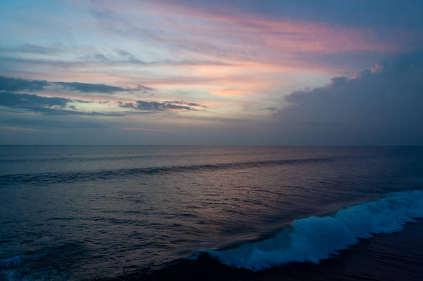 A peaceful seascape during golden hour featuring a soft pink and blue sunset sky over the calm deep blue ocean with a gentle white wave breaking in the foreground.