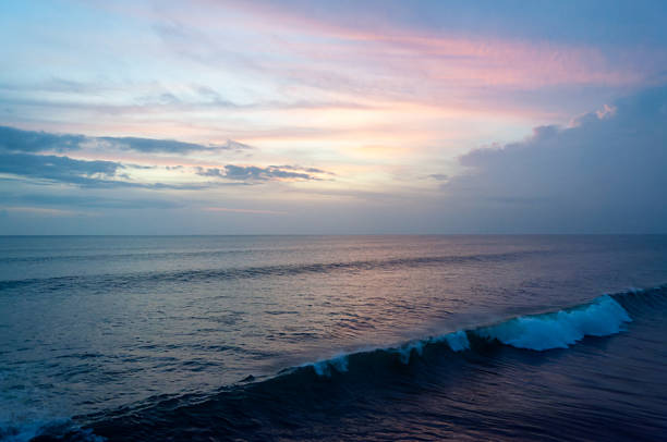 A peaceful seascape during golden hour featuring a soft pink and blue sunset sky over the calm deep blue ocean with a gentle white wave breaking in the foreground.