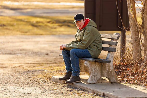 In the warm glow of an early spring afternoon in New Jersey, a young man sits quietly on a stone bench, absorbed in thought. Dressed in a green jacket, blue jeans, and sturdy boots, he gazes thoughtfully into the distance. His red hoodie peeks out from under his jacket, adding a splash of color to the serene scene. The park around him is tranquil, with bare trees and a soft golden light that suggests the season is just beginning to change.